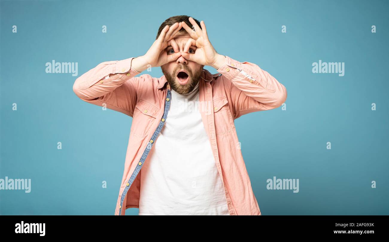 Strange bearded man makes a gesture like binoculars, pressing his hands to face, eyes look through fingers and is surprised. Stock Photo