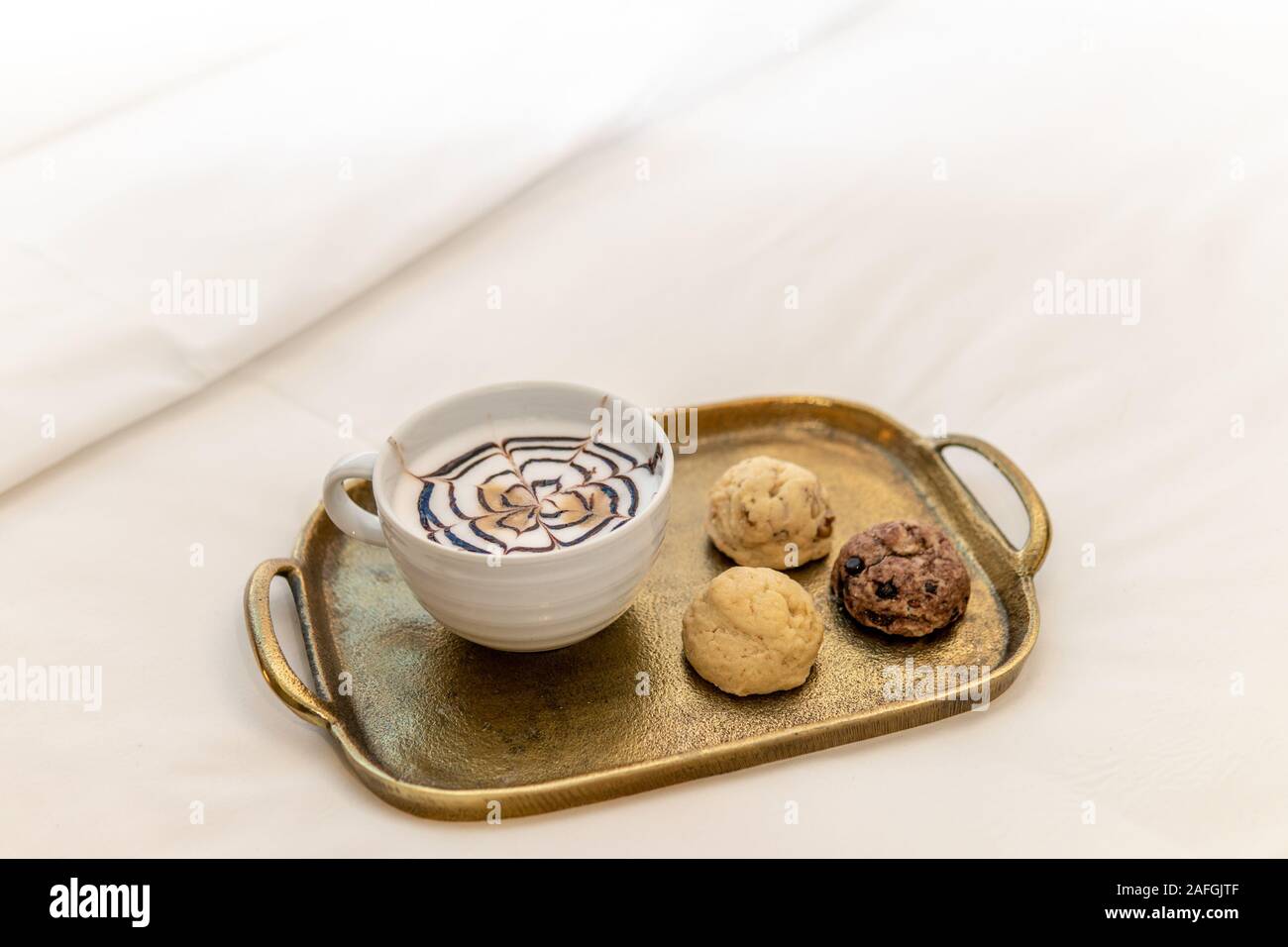 cappuccino with fancy chocolate design on top with three various baked cookies on gold tray in hotel room on bed Stock Photo