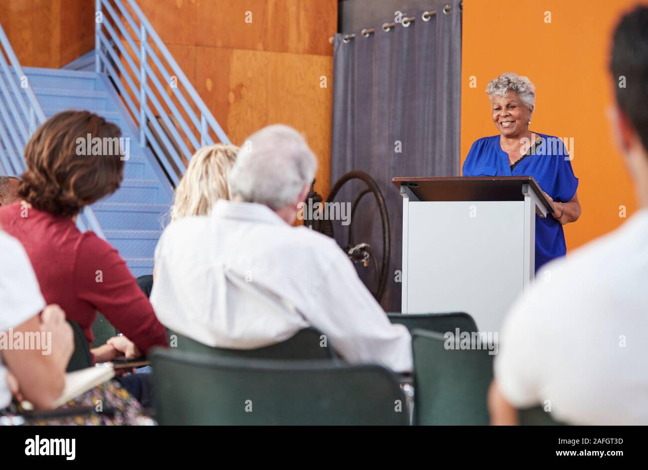 Senior Woman At Podium Chairing Neighborhood Meeting In Community Centre Stock Photo