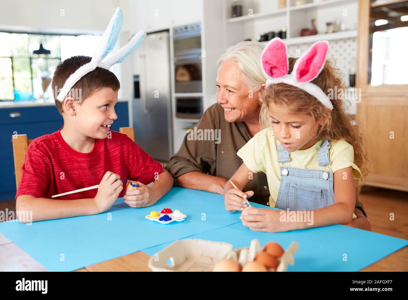 Grandmother With Grandchildren Wearing Rabbit Ears Decorating Easter Eggs At Home Together Stock Photo