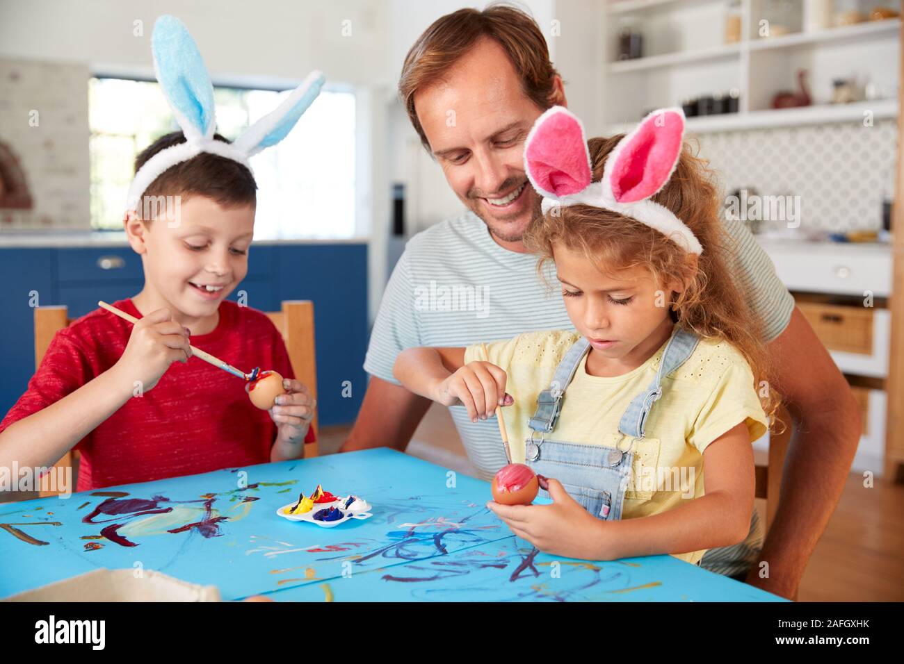 Father With Children Wearing Rabbit Ears Decorating Easter Eggs At Home Together Stock Photo