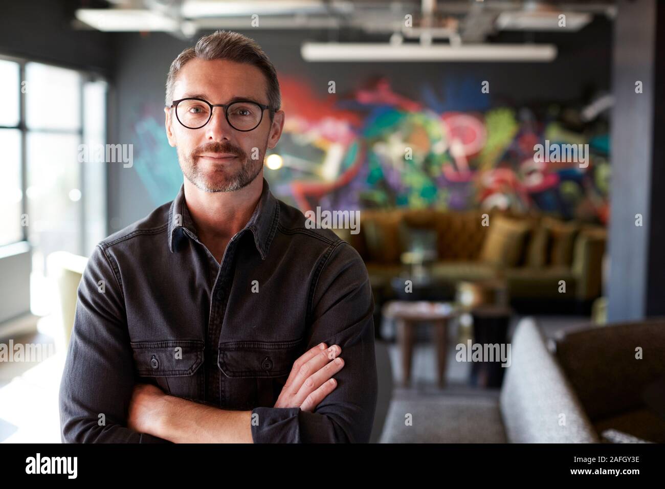 Middle aged white male creative in casual office lounge area looks to camera, arms crossed, waist up Stock Photo