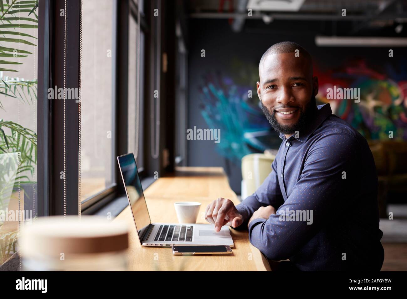Mid adult black male creative sitting by window in cafe using laptop, turning and smiling to camera Stock Photo