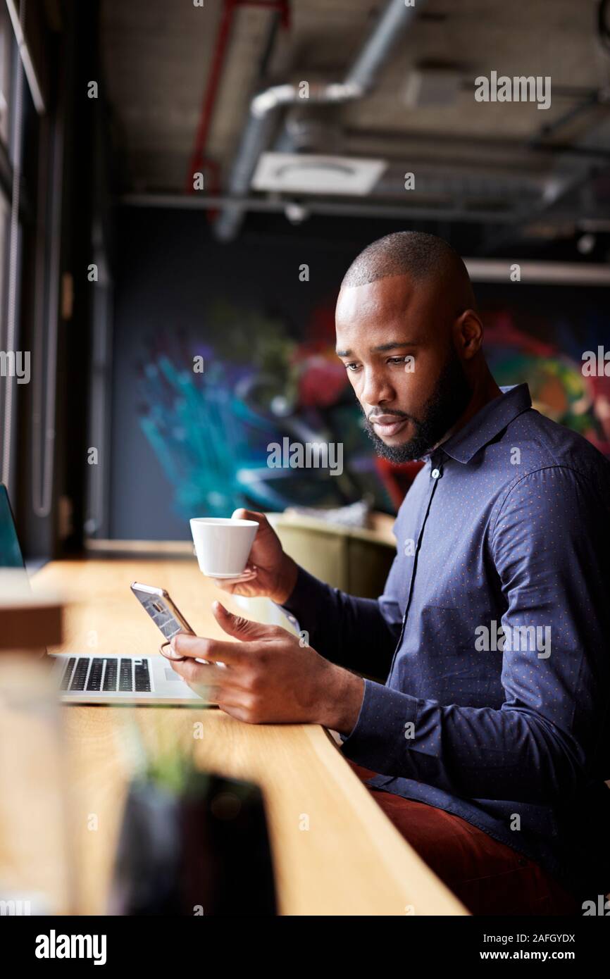 Mid adult black male creative sitting by window having coffee using smartphone, side view, vertical Stock Photo