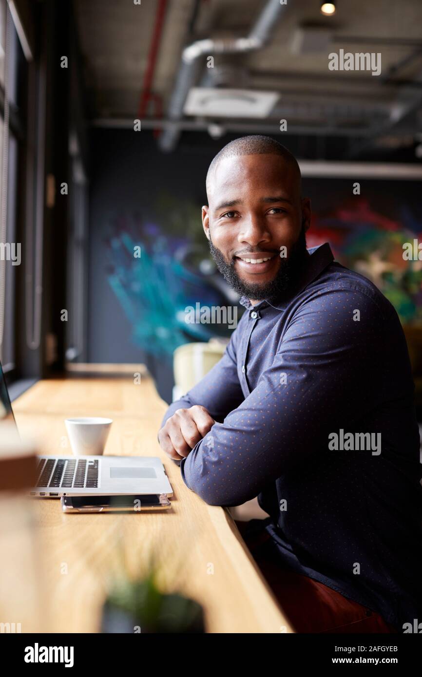 Black male creative sitting by window in cafe using laptop, turning and smiling to camera, vertical Stock Photo