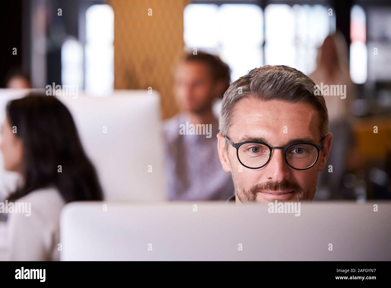 Middle aged white male creative using a computer in a busy office, selective focus, close up Stock Photo