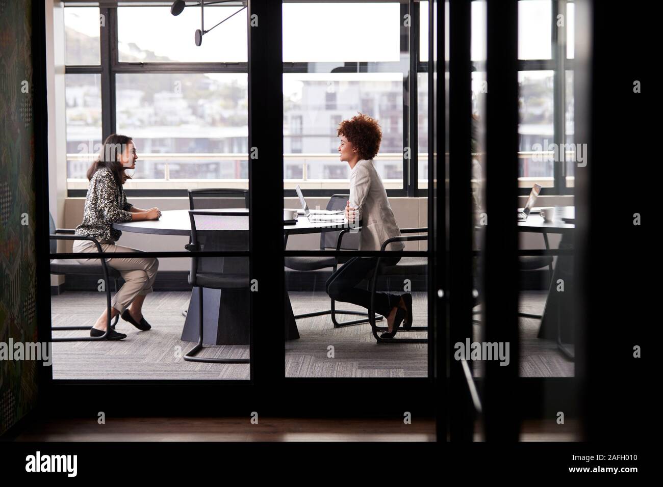Two millennial businesswomen meeting for a job interview, full length, seen through glass wall Stock Photo