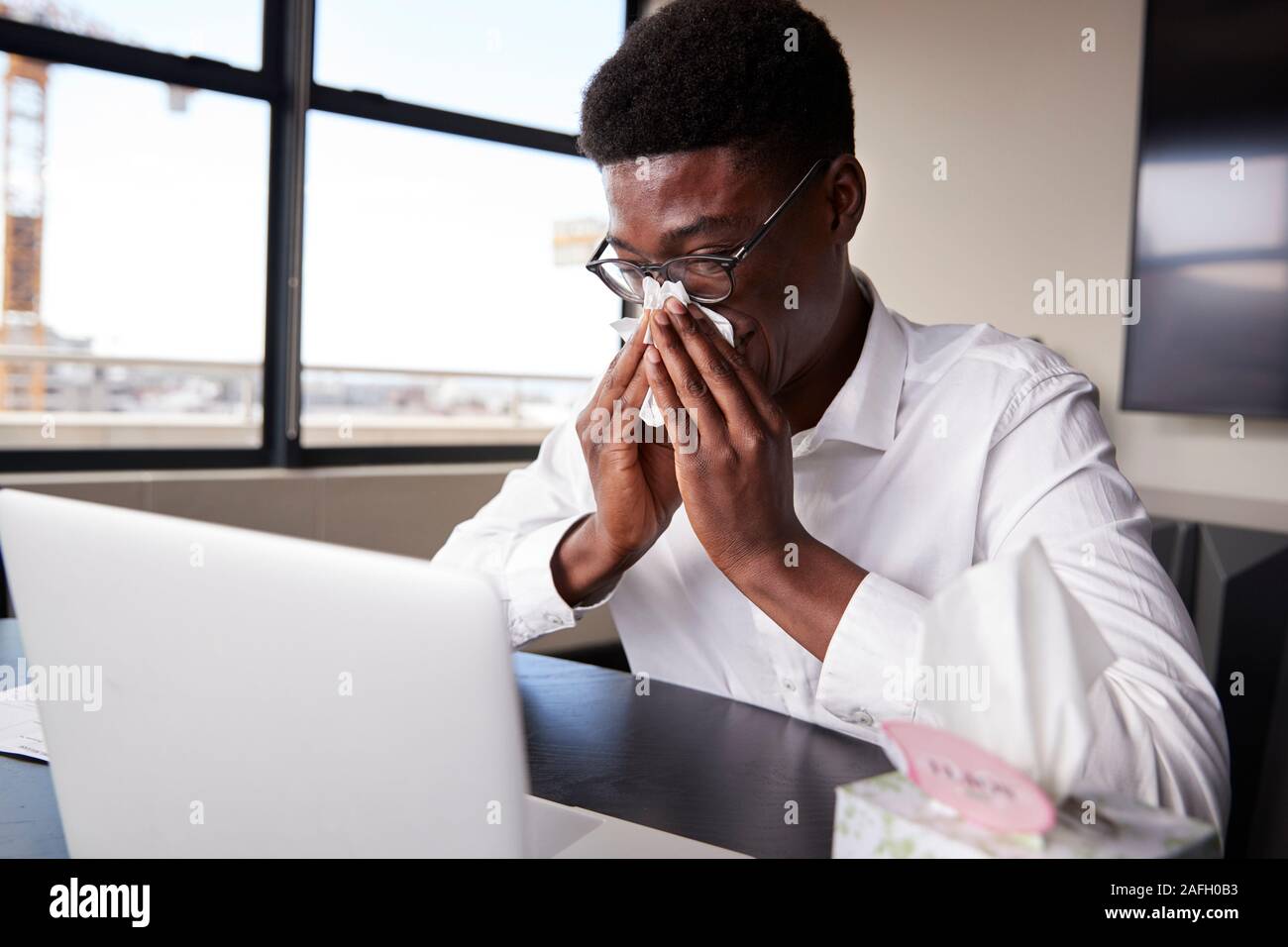 Young black businessman sitting at an office desk blowing his nose into a tissue, close up Stock Photo