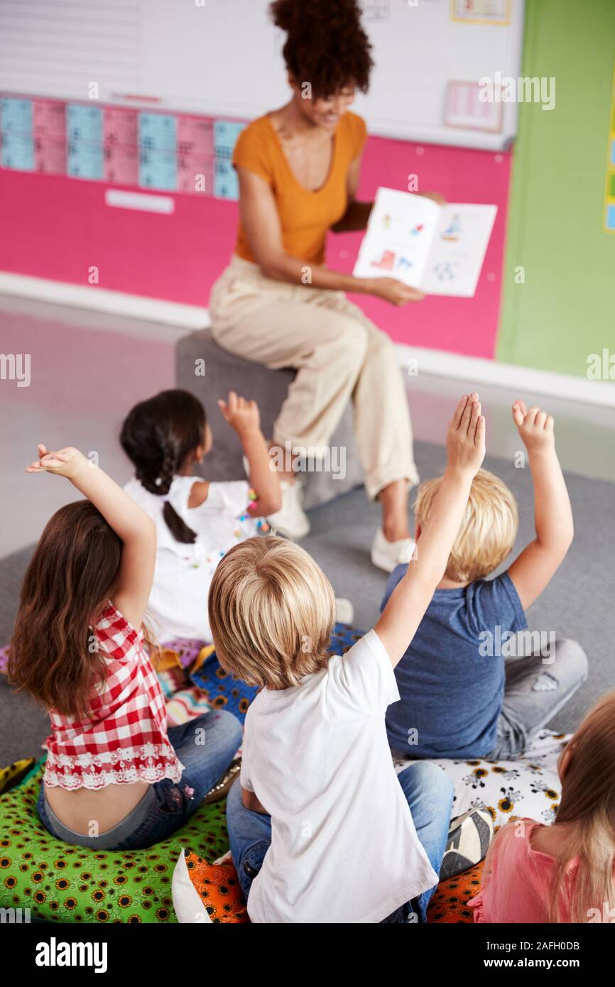 Elementary Pupils Raising Hands To Answer Question As Female Teacher Reads Story In Classroom Stock Photo