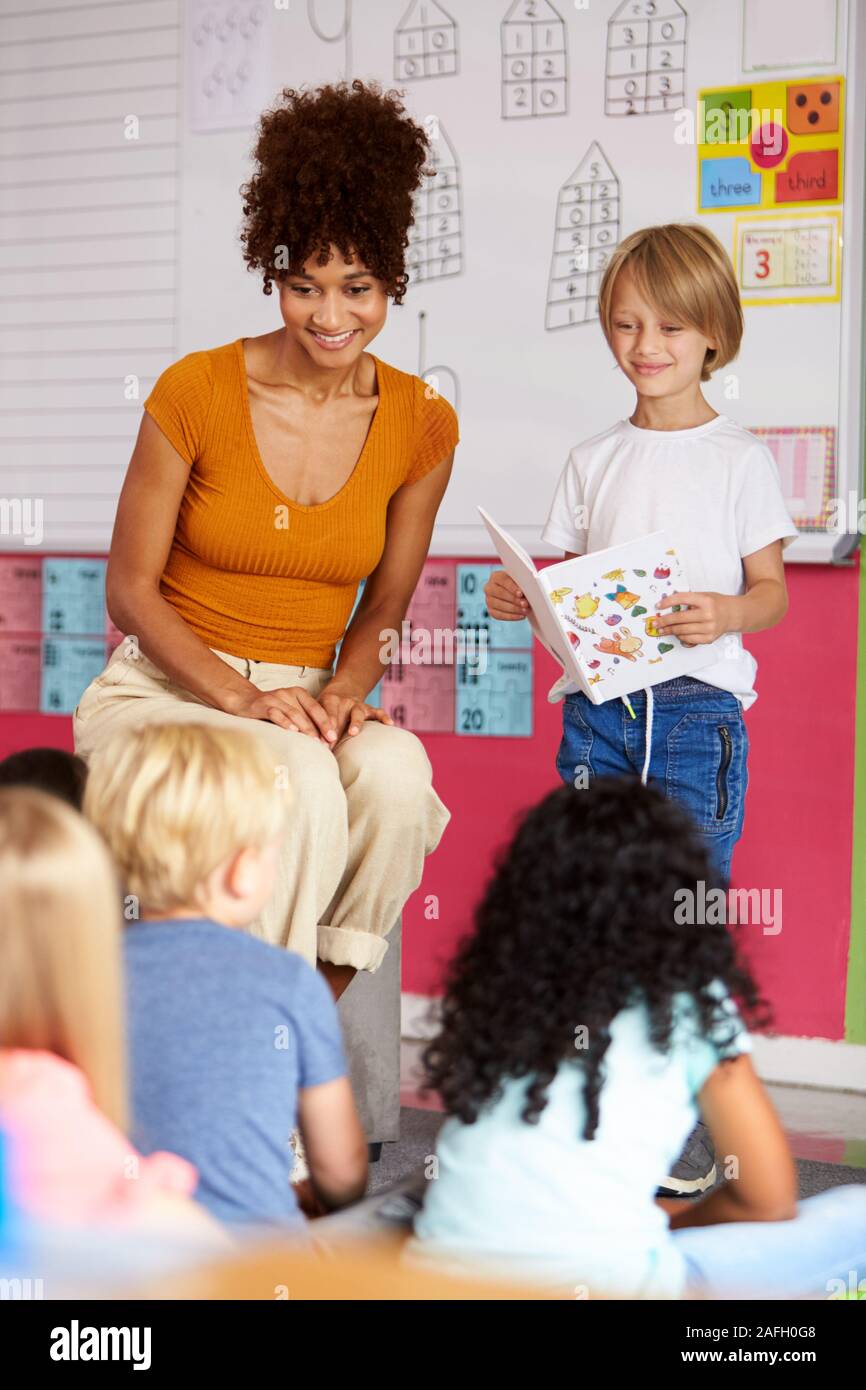 Male Pupil In Elementary School Classroom Reading Book To Class With Teacher Stock Photo