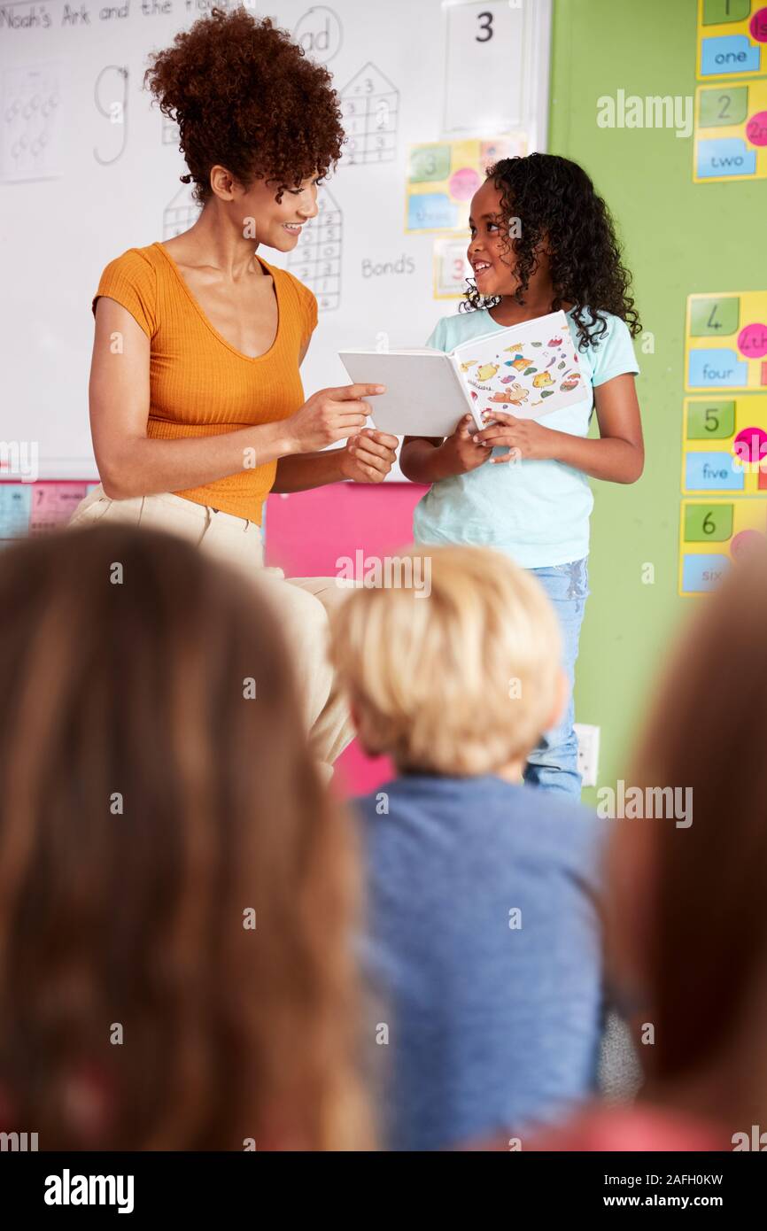 Female Pupil In Elementary School Classroom Reading Book To Class With Teacher Stock Photo