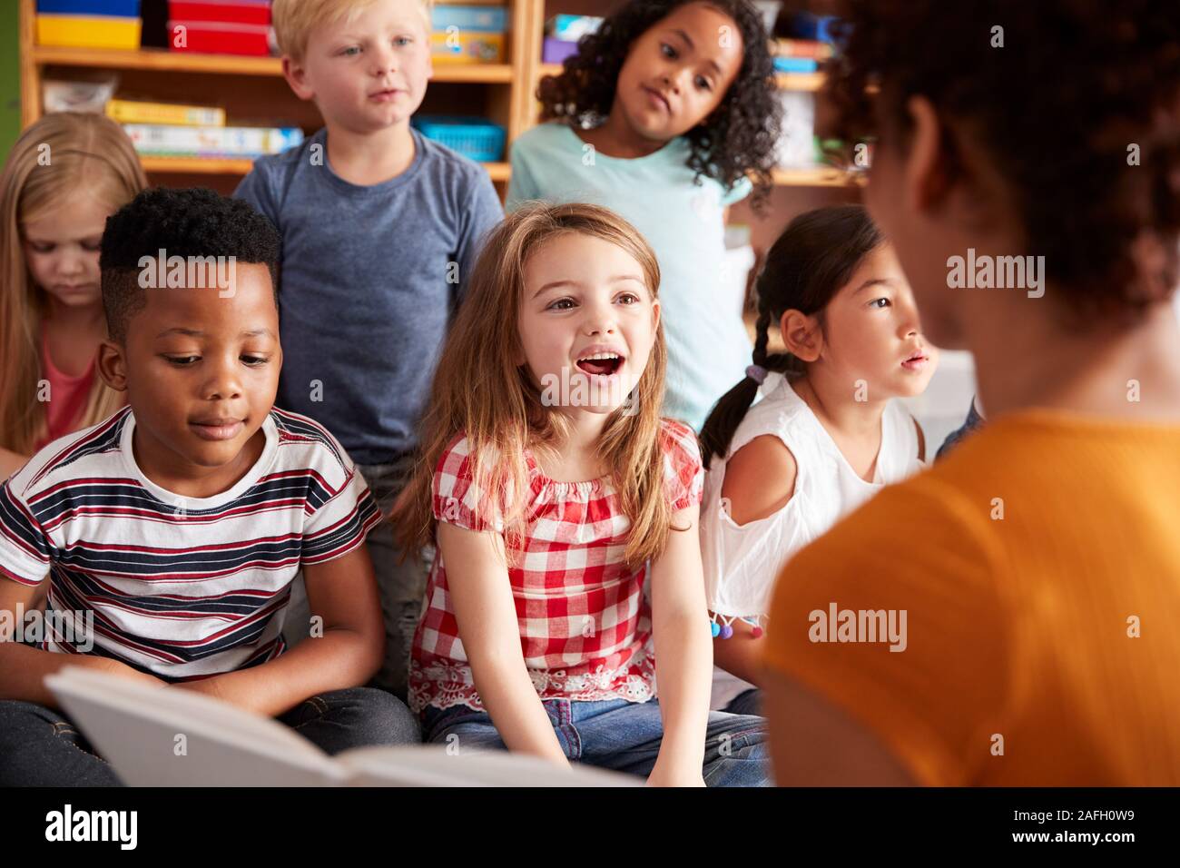 Group Of Elementary School Pupils Sitting On Floor Listening To Female Teacher Read Story Stock Photo
