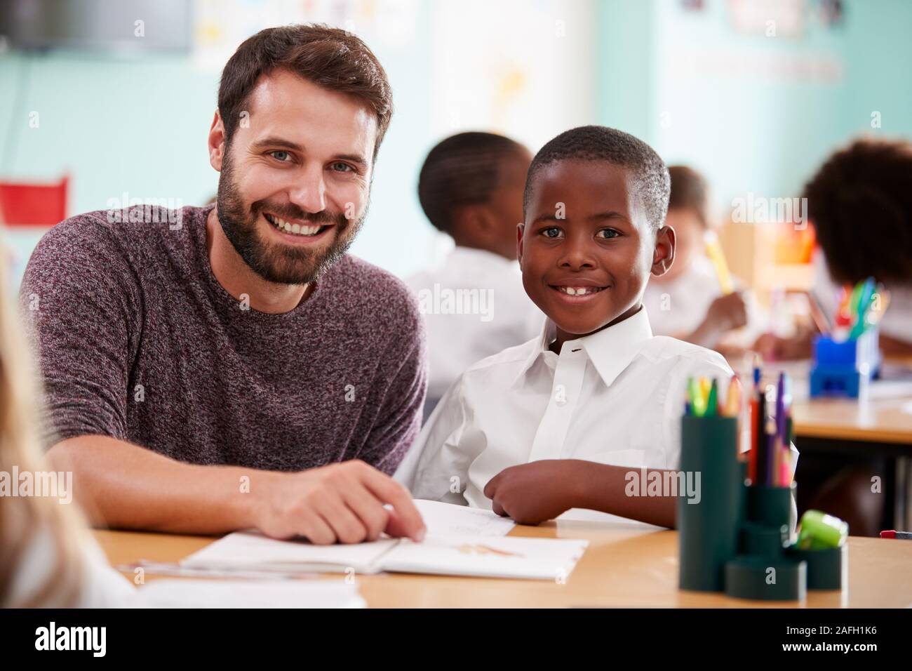 Portrait Of Elementary School Teacher Giving Male Pupil Wearing Uniform One To One Support In Class Stock Photo
