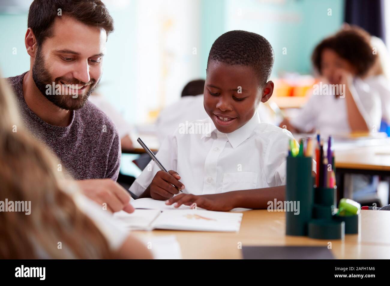 Elementary School Teacher Giving Male Pupil Wearing Uniform One To One Support In Classroom Stock Photo