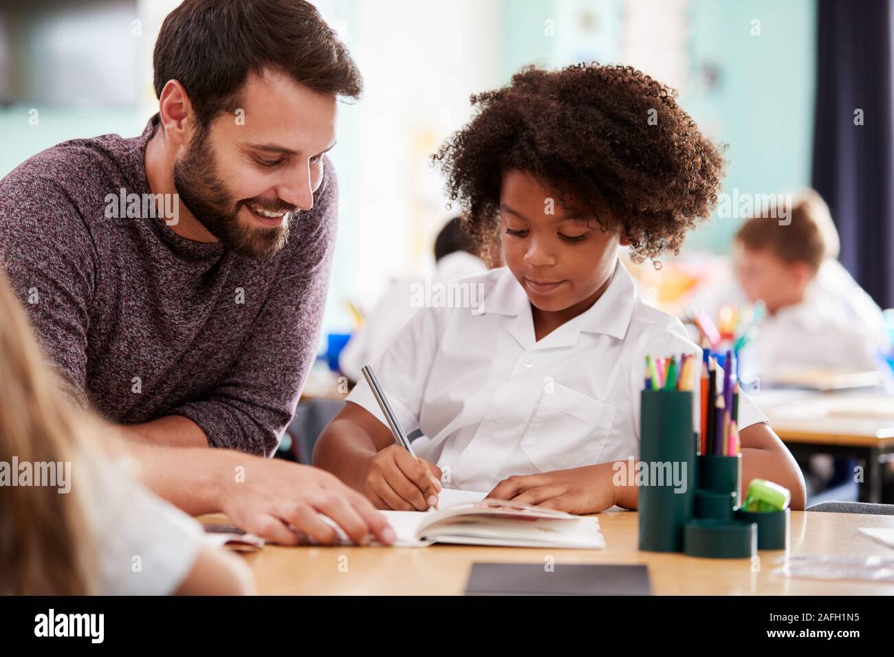 Male Elementary School Teacher Giving Female Pupil Wearing Uniform One To One Support In Classroom Stock Photo