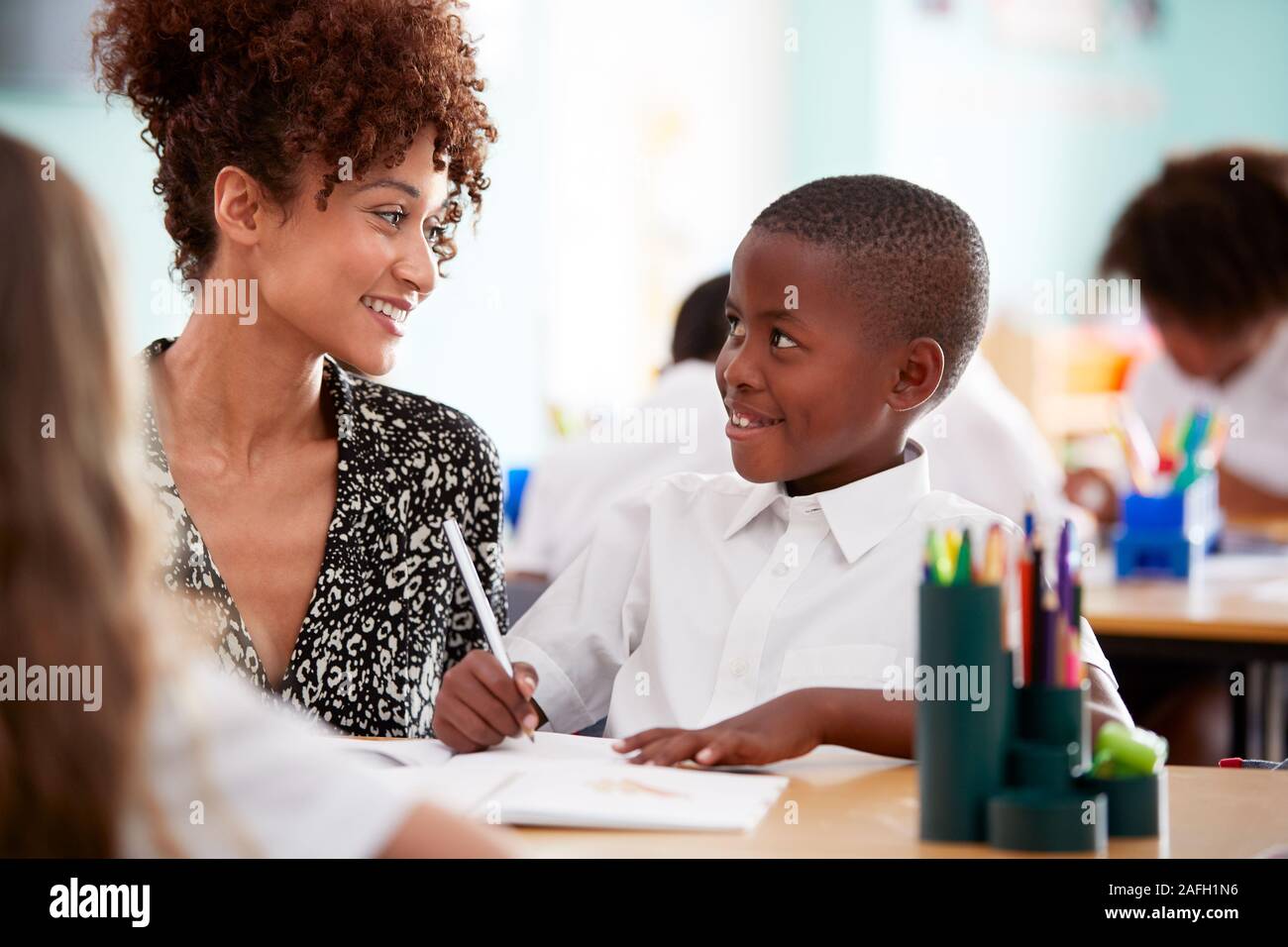 Woman Elementary School Teacher Giving Male Pupil Wearing Uniform One To One Support In Classroom Stock Photo