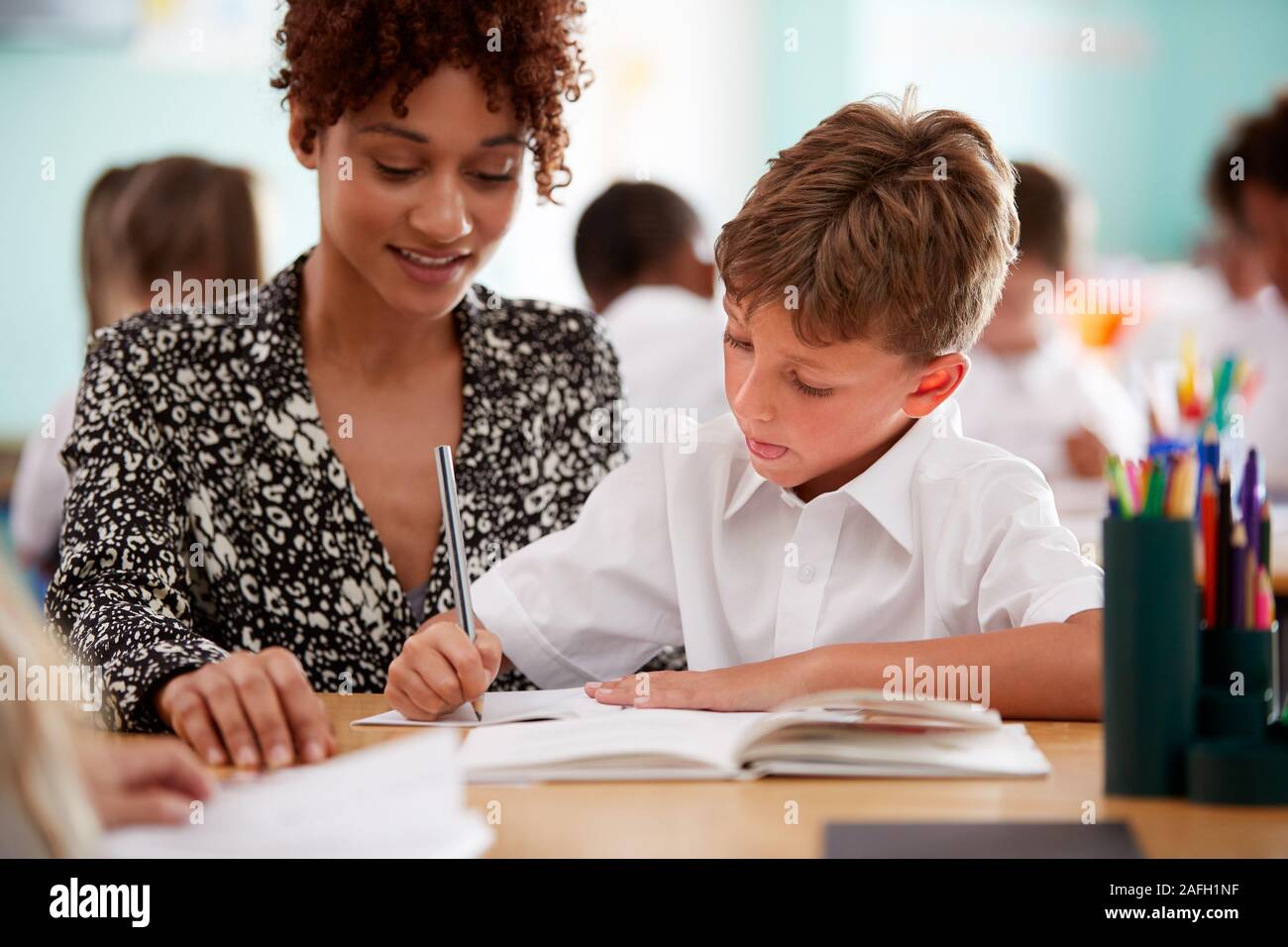 Woman Elementary School Teacher Giving Male Pupil Wearing Uniform One To One Support In Classroom Stock Photo