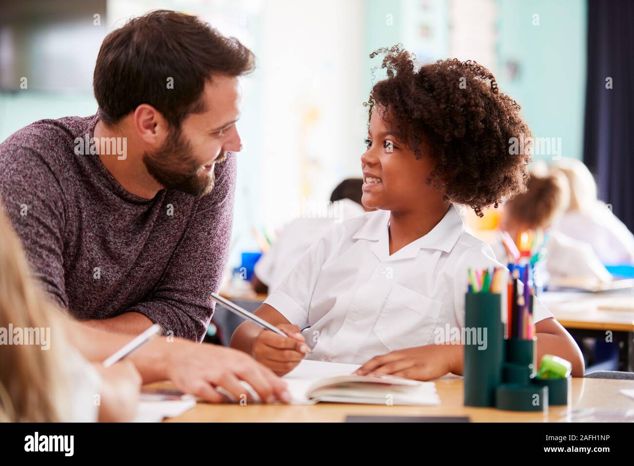 Male Elementary School Teacher Giving Female Pupil Wearing Uniform One To One Support In Classroom Stock Photo