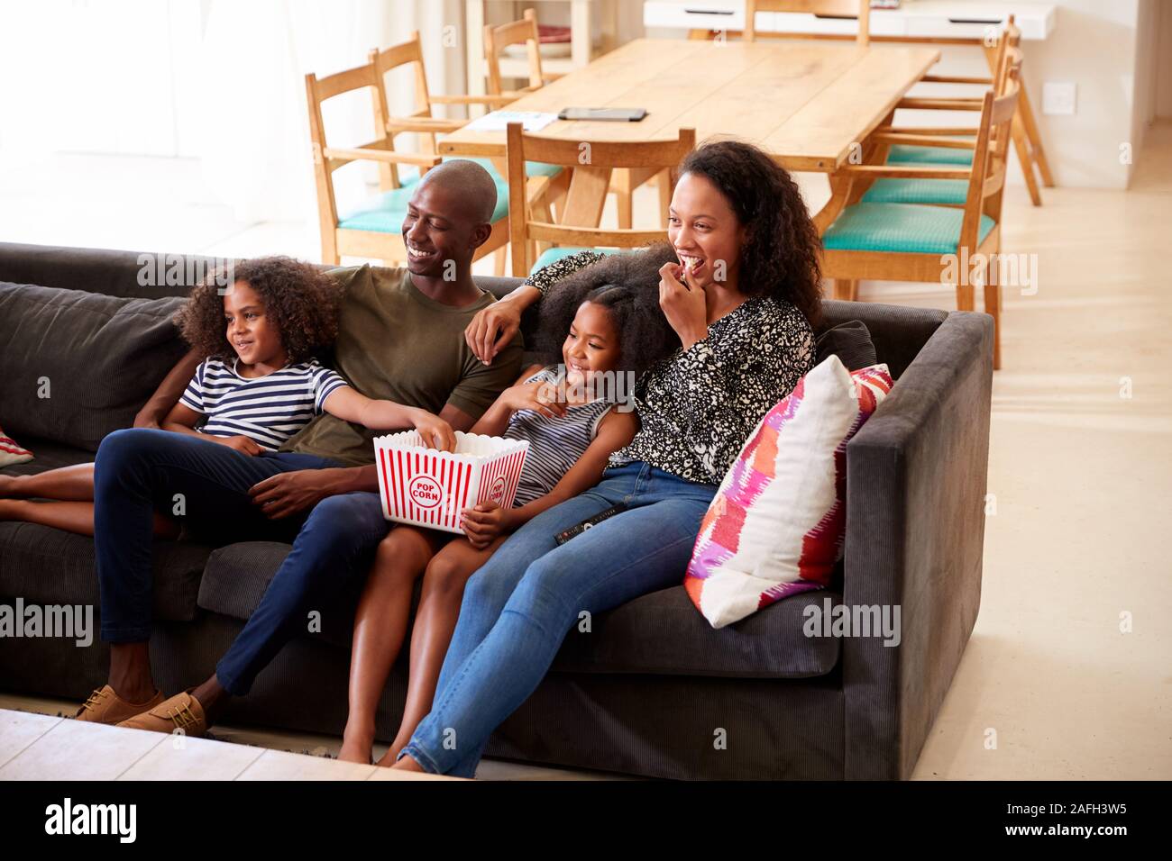 Family Sitting On Sofa At Home Eating Popcorn And Watching Movie Together Stock Photo