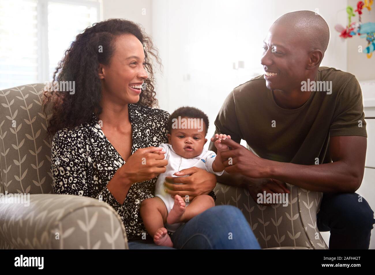 Loving Parents Sitting In Chair Cuddling Baby Son In Nursery At Home Stock Photo