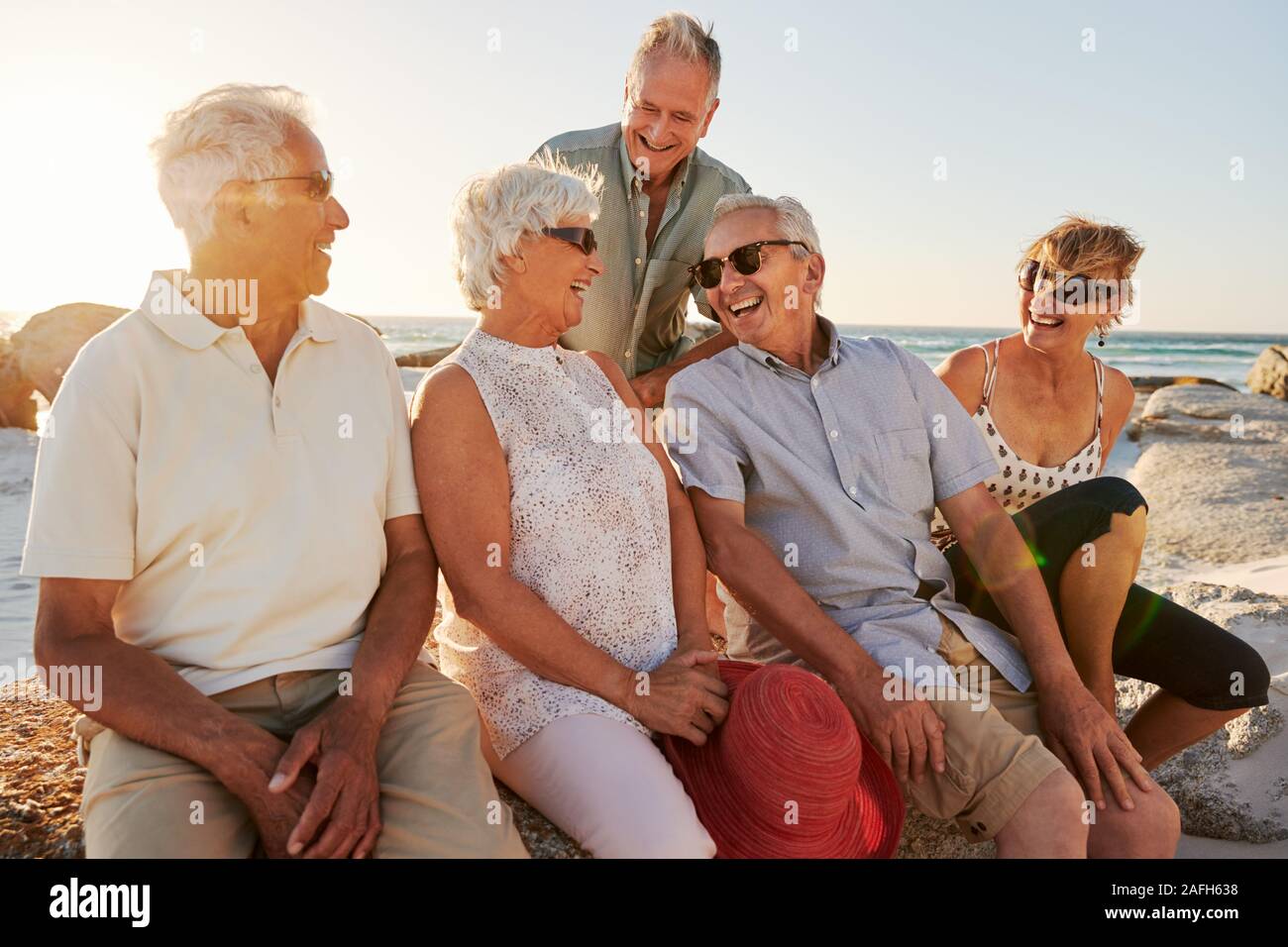 Group Of Senior Friends Sitting On Rocks By Sea On Summer Group Vacation Stock Photo