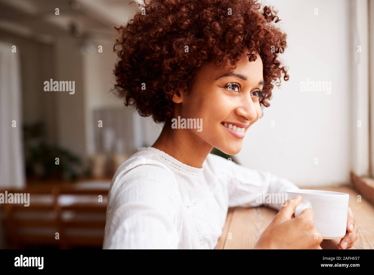 Young Woman Relaxing In Loft Apartment Looking Out Of Window With Hot Drink Stock Photo