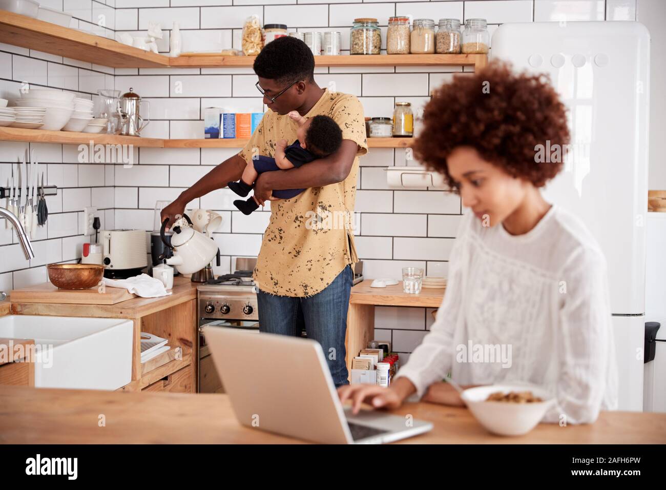 Multi-Tasking Father Holds Baby Son And Pours Drink As Mother Uses Laptop And Eats Breakfast Stock Photo