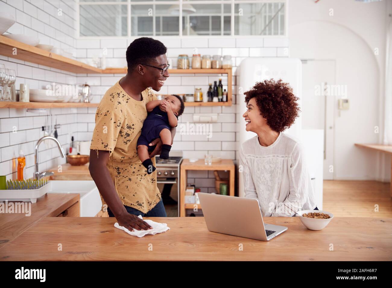 Multi-Tasking Father Holds Baby Son And Cleans Surface As Mother Uses Laptop And Eats Breakfast Stock Photo