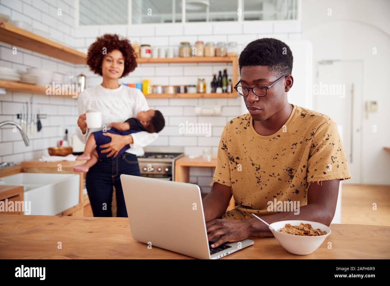 Multi-Tasking Mother Holds Baby Son And Makes Hot Drink As Father Uses Laptop And Eats Breakfast Stock Photo
