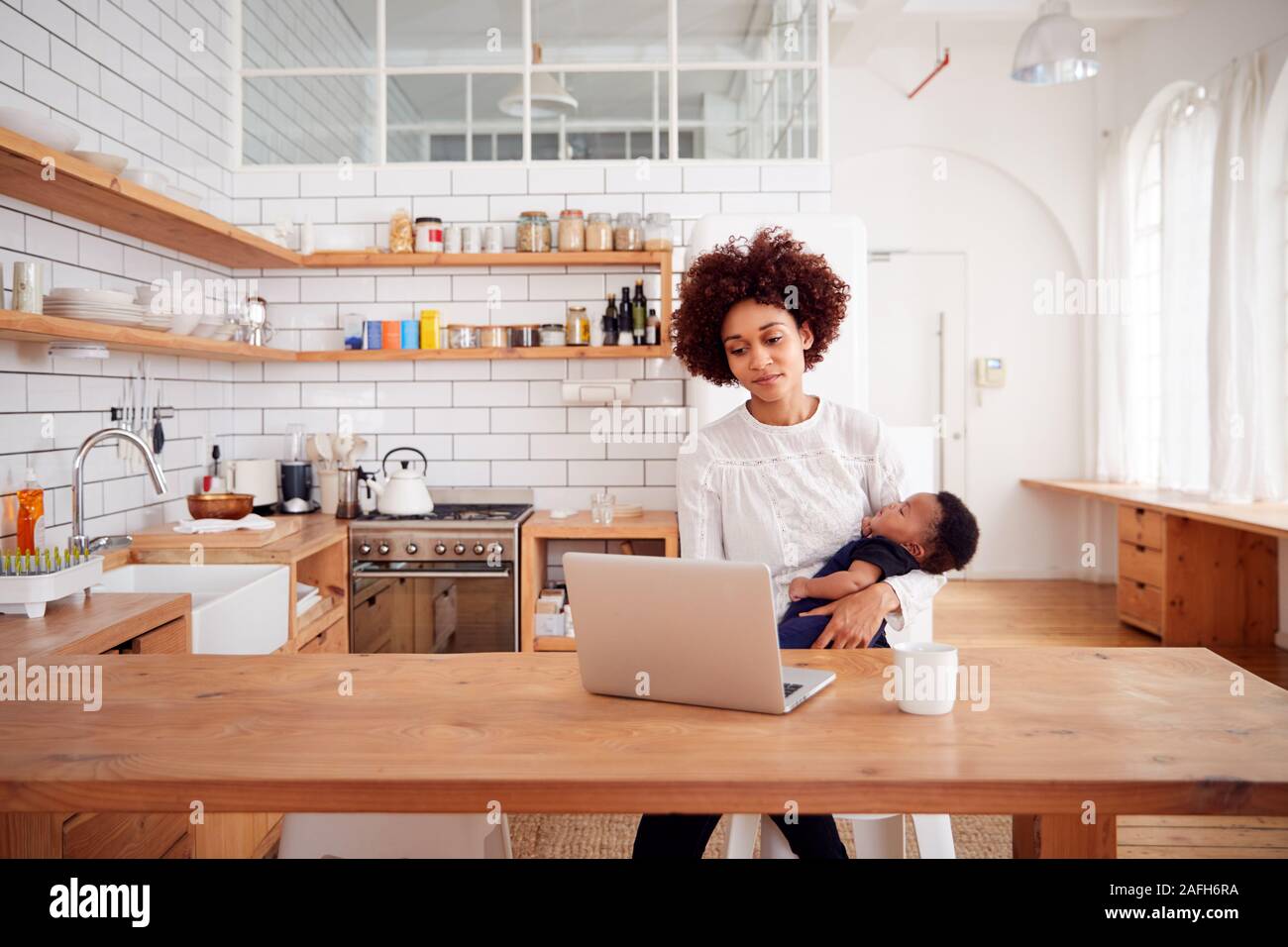 Multi-Tasking Mother Holds Sleeping Baby Son And Works On Laptop Computer In Kitchen Stock Photo