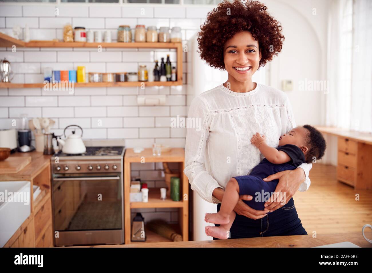 Portrait Of Smiling Mother Holding Sleeping Baby Son In Kitchen Stock Photo