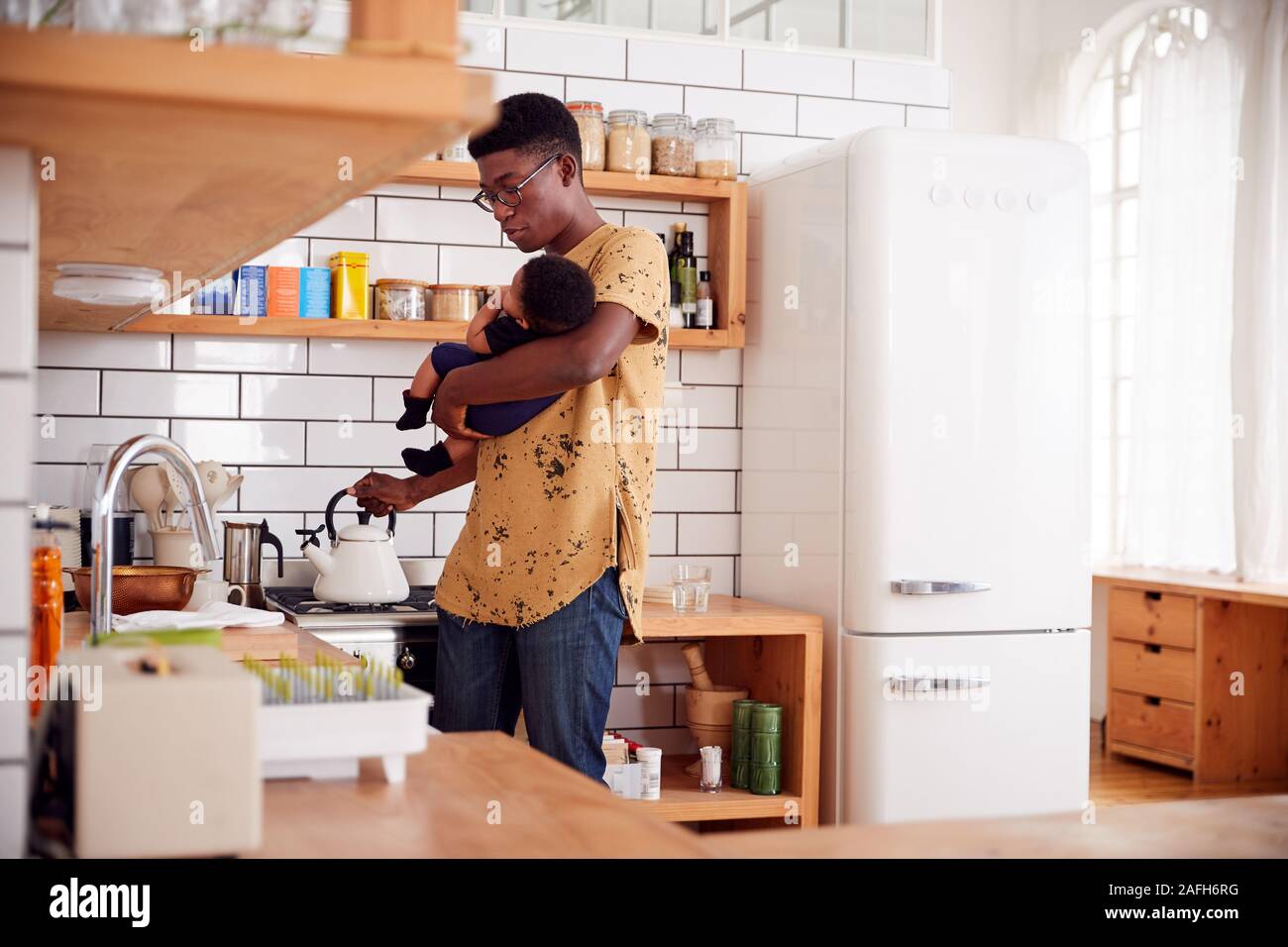 Multi-Tasking Father Holds Baby Son And Pours Kettle In Kitchen Stock Photo