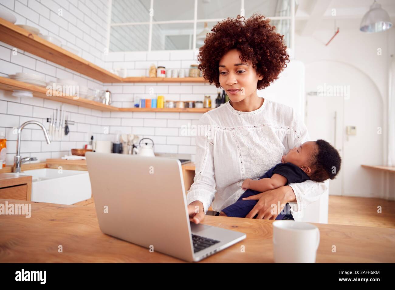 Multi-Tasking Mother Holds Sleeping Baby Son And Works On Laptop Computer In Kitchen Stock Photo