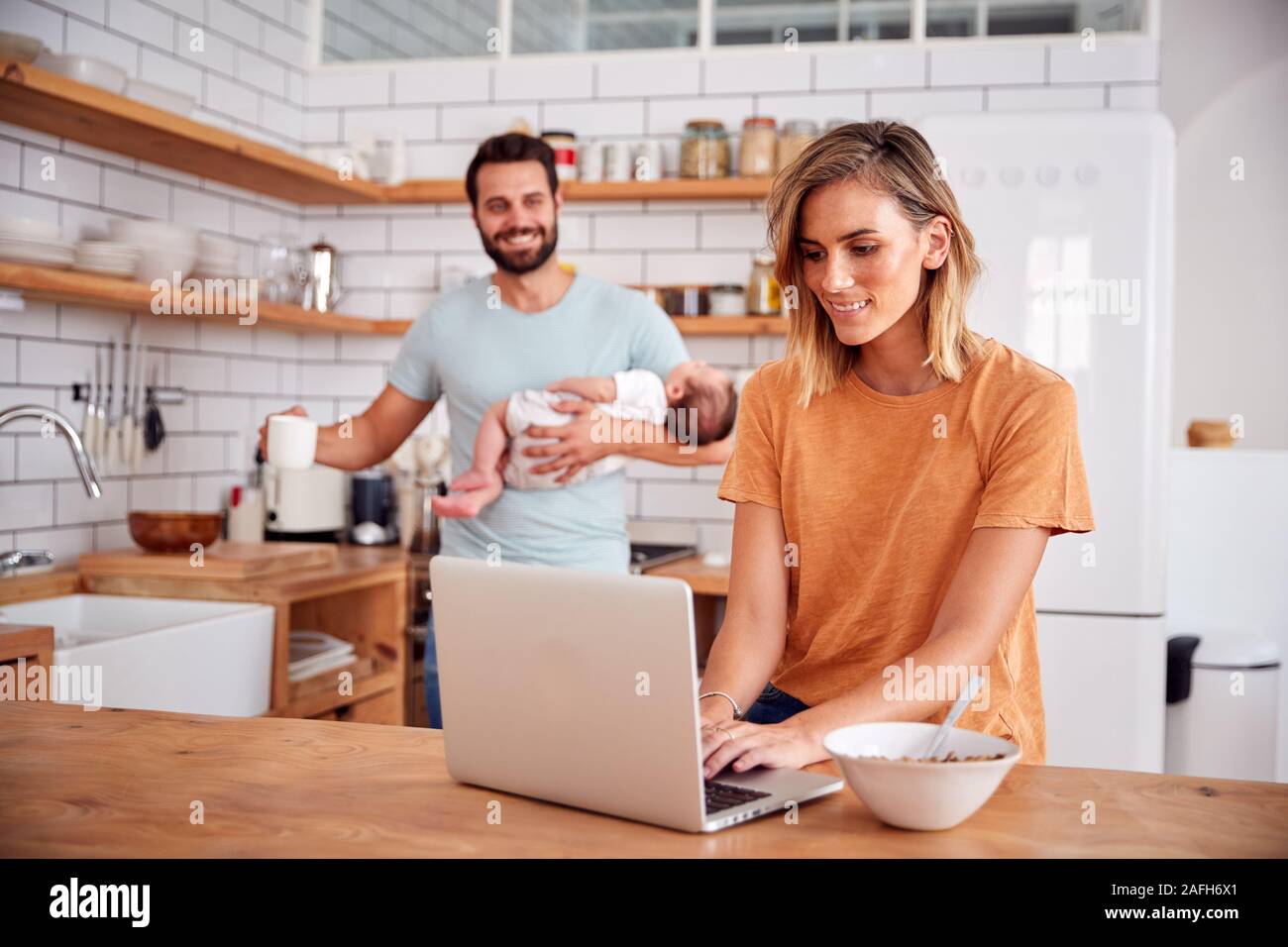 Multi-Tasking Father Holds Baby Son And Makes Hot Drink As Mother Uses Laptop And Eats Breakfast Stock Photo