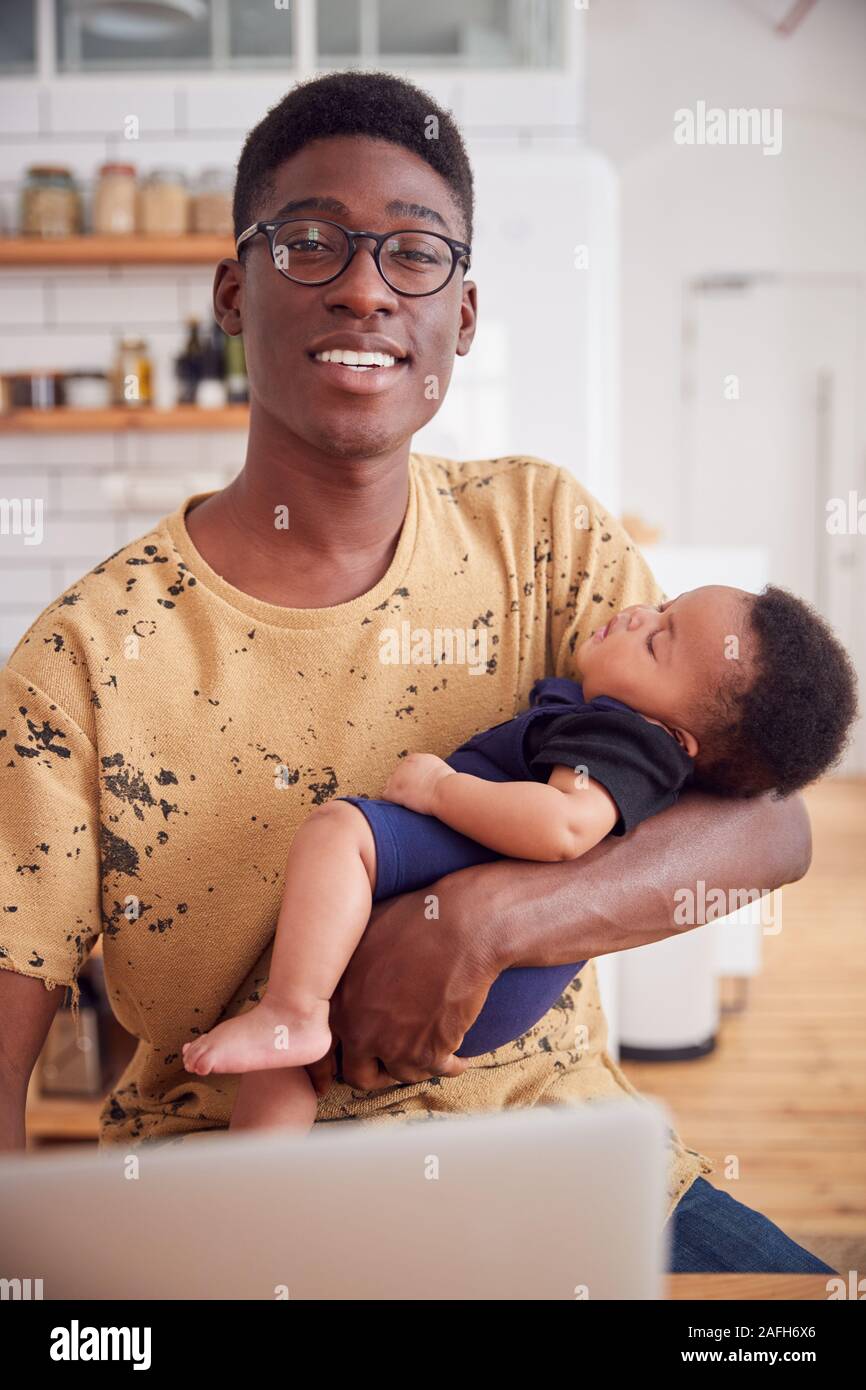 Portrait Of Multi-Tasking Father Holding Sleeping Baby Son And Working On Laptop Computer In Kitchen Stock Photo