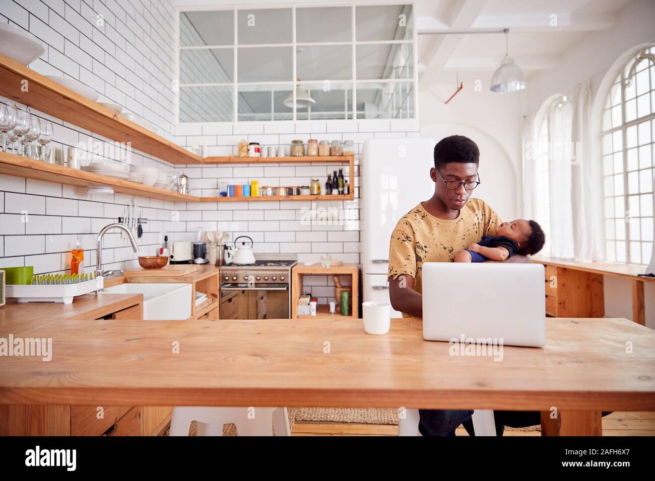 Multi-Tasking Father Holds Sleeping Baby Son And Works On Laptop Computer In Kitchen Stock Photo