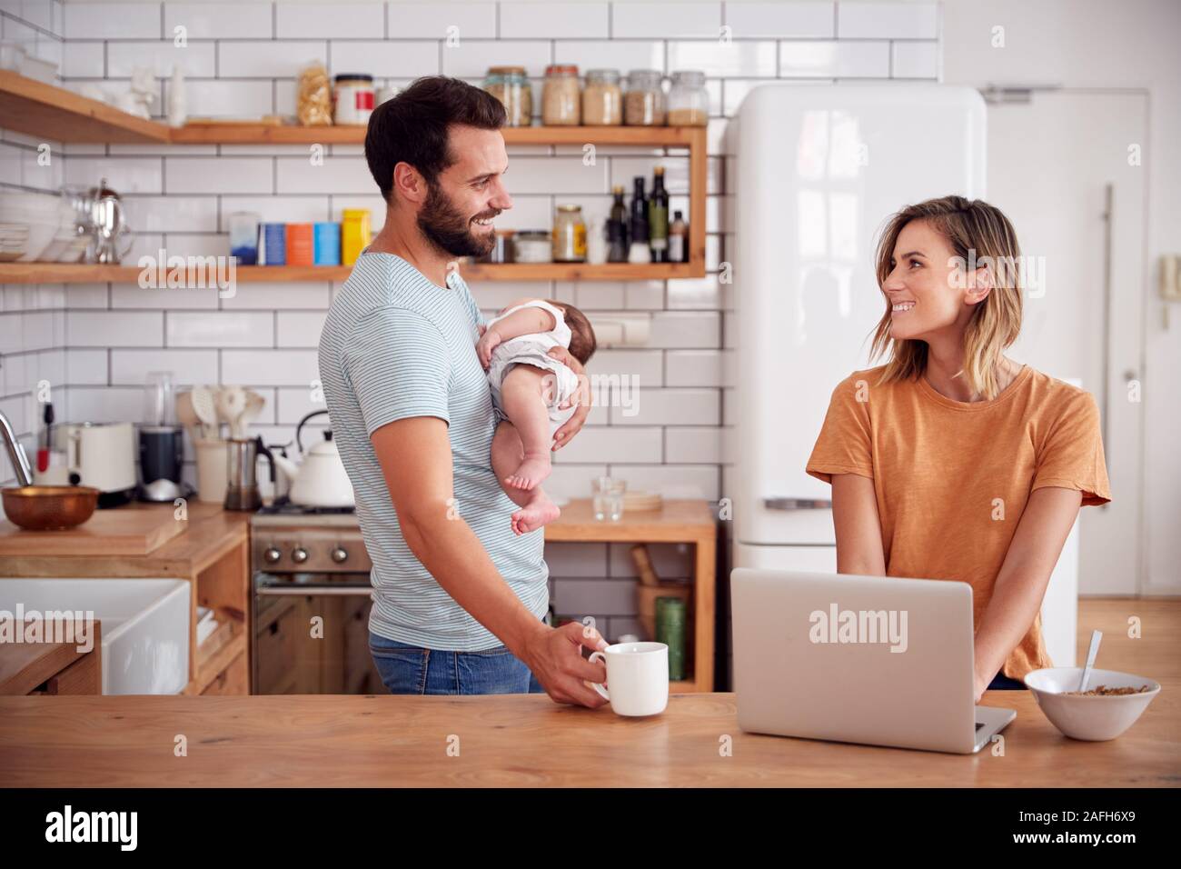 Multi-Tasking Father Holds Baby Son And Makes Hot Drink As Mother Uses Laptop And Eats Breakfast Stock Photo