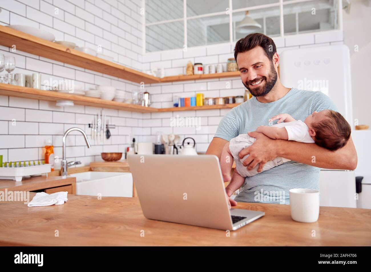 Multi-Tasking Father Holds Sleeping Baby Son And Works On Laptop Computer In Kitchen Stock Photo