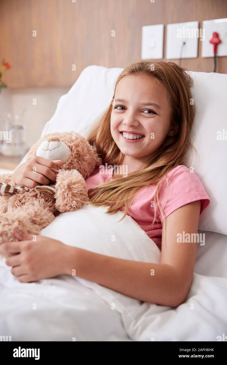 Portrait Of Girl Lying In Bed In Hospital Ward Hugging Teddy Bear Stock