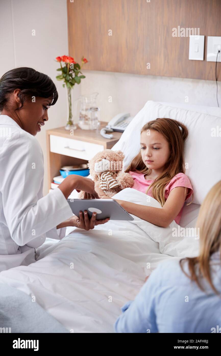 Female Doctor Visiting Mother And Daughter Lying In Bed In Hospital Ward Using Digital Tablet Stock Photo