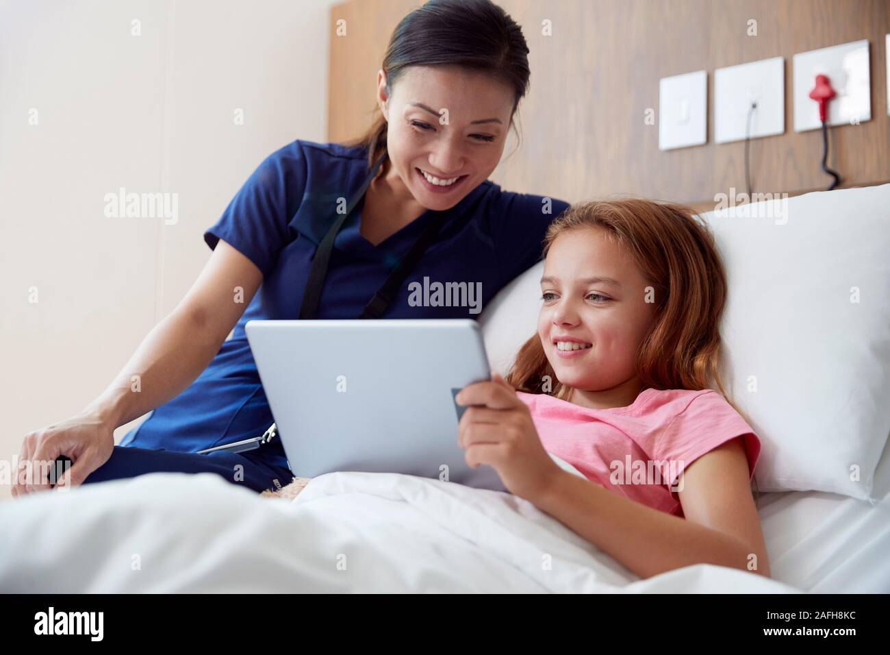 Female Nurse With Girl Lying In Hospital Bed Looking At Digital Tablet Together Stock Photo