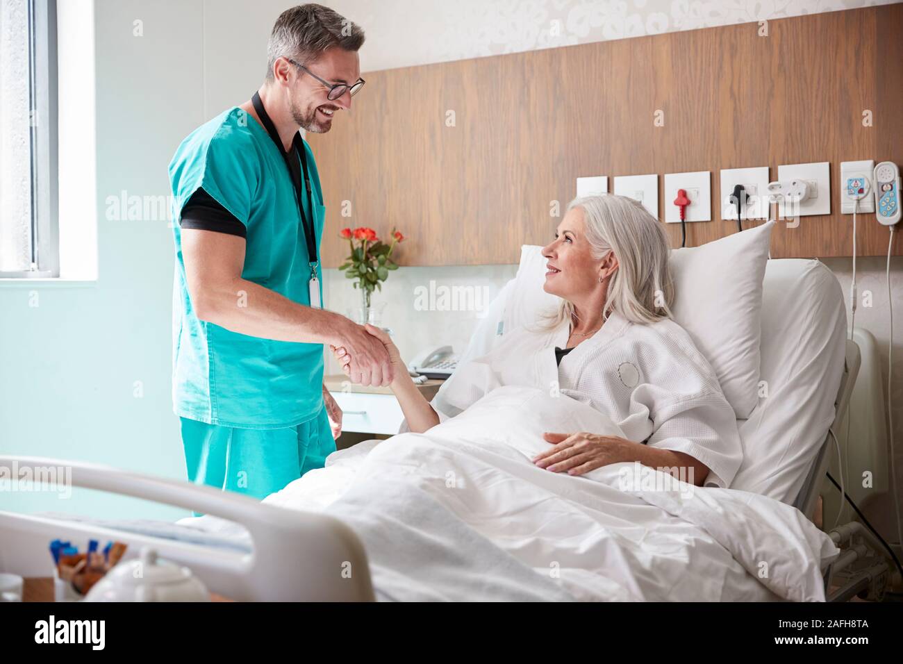 Surgeon Visiting And Shaking Hands With Mature Female Patient In Hospital Bed Stock Photo