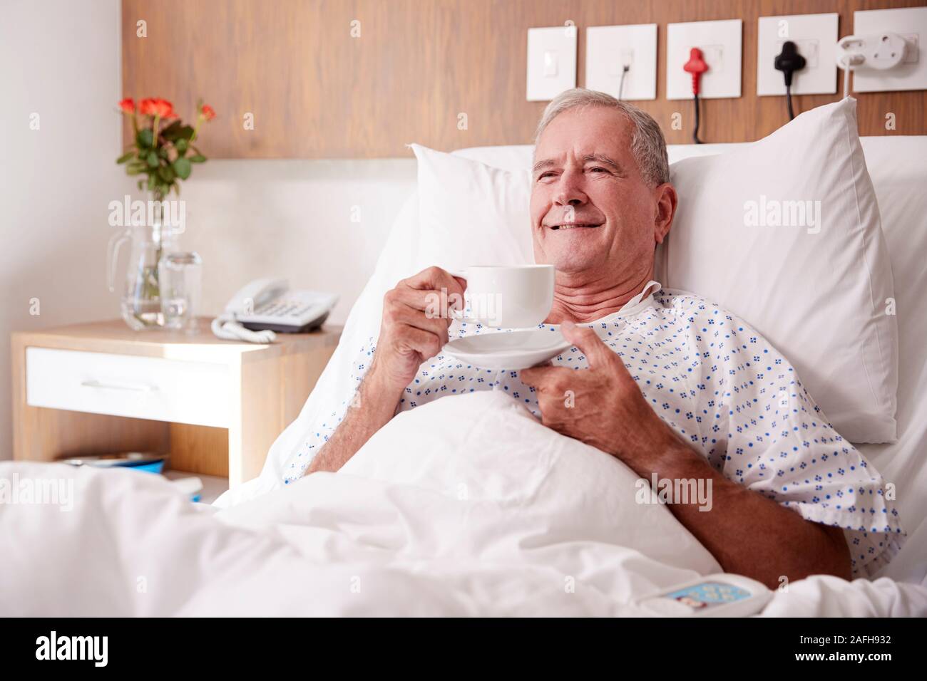 Male Senior Patient Lying In Hospital Bed Enjoying Hot Drink Stock Photo