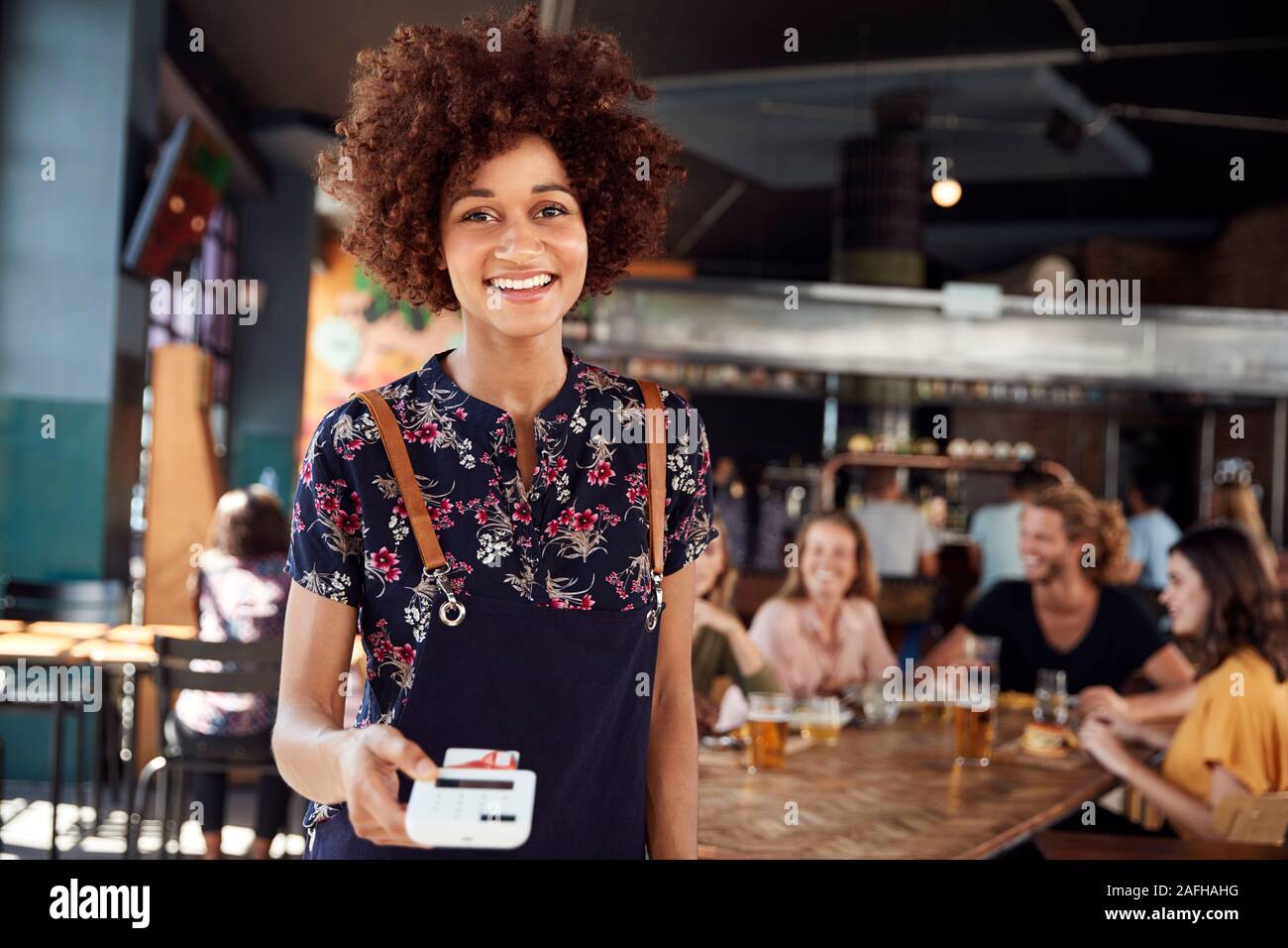Portrait Of Waitress Holding Credit Card Payment Terminal In Busy Bar Restaurant Stock Photo