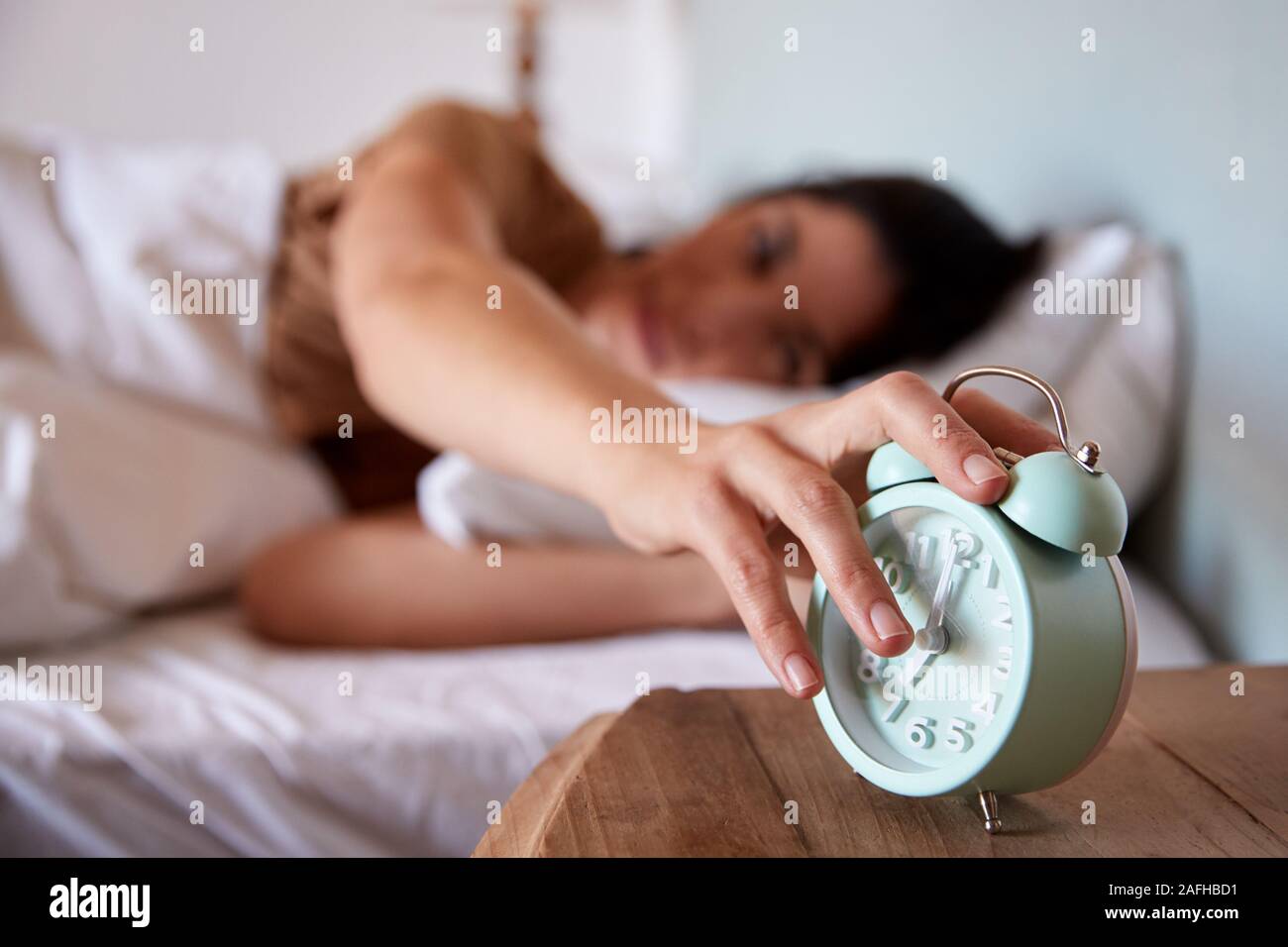 Mid adult woman lying in bed, reaching out to alarm clock on the bedside table in the foreground Stock Photo