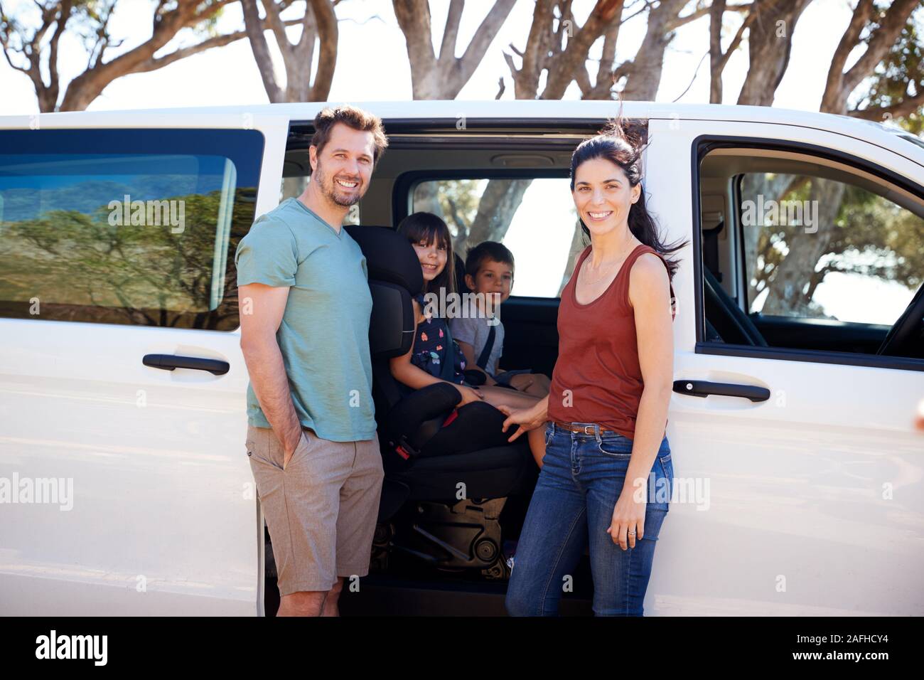 Mid adult white parents standing beside car, their two young kids inside smiling to camera Stock Photo