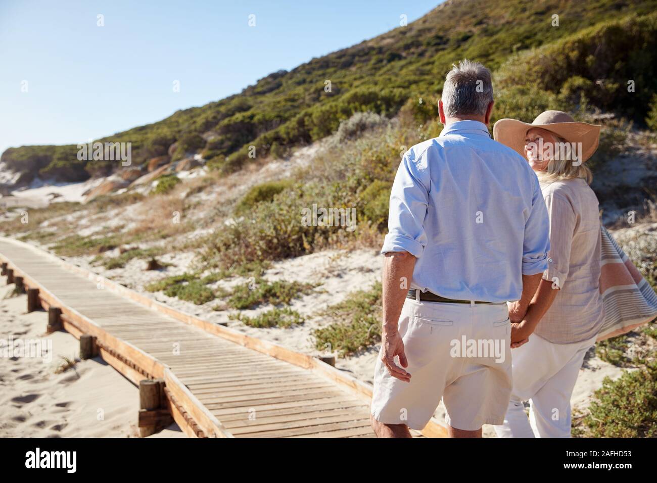 Senior white couple walking along a wooden promenade on a beach holding hands, close up, back view Stock Photo