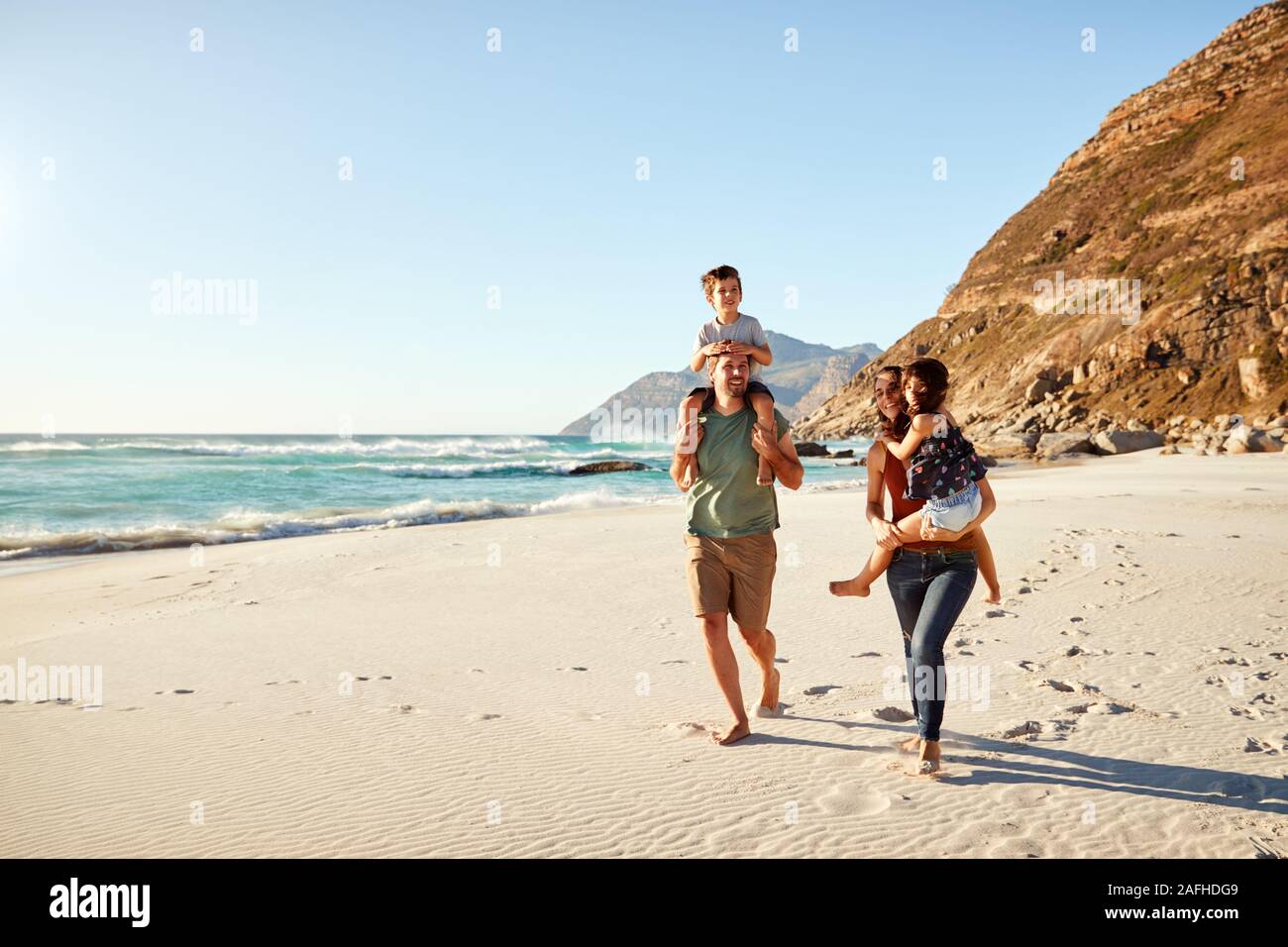 Mid adult white couple walking along on a beach on a family holiday carrying their kids, full length Stock Photo