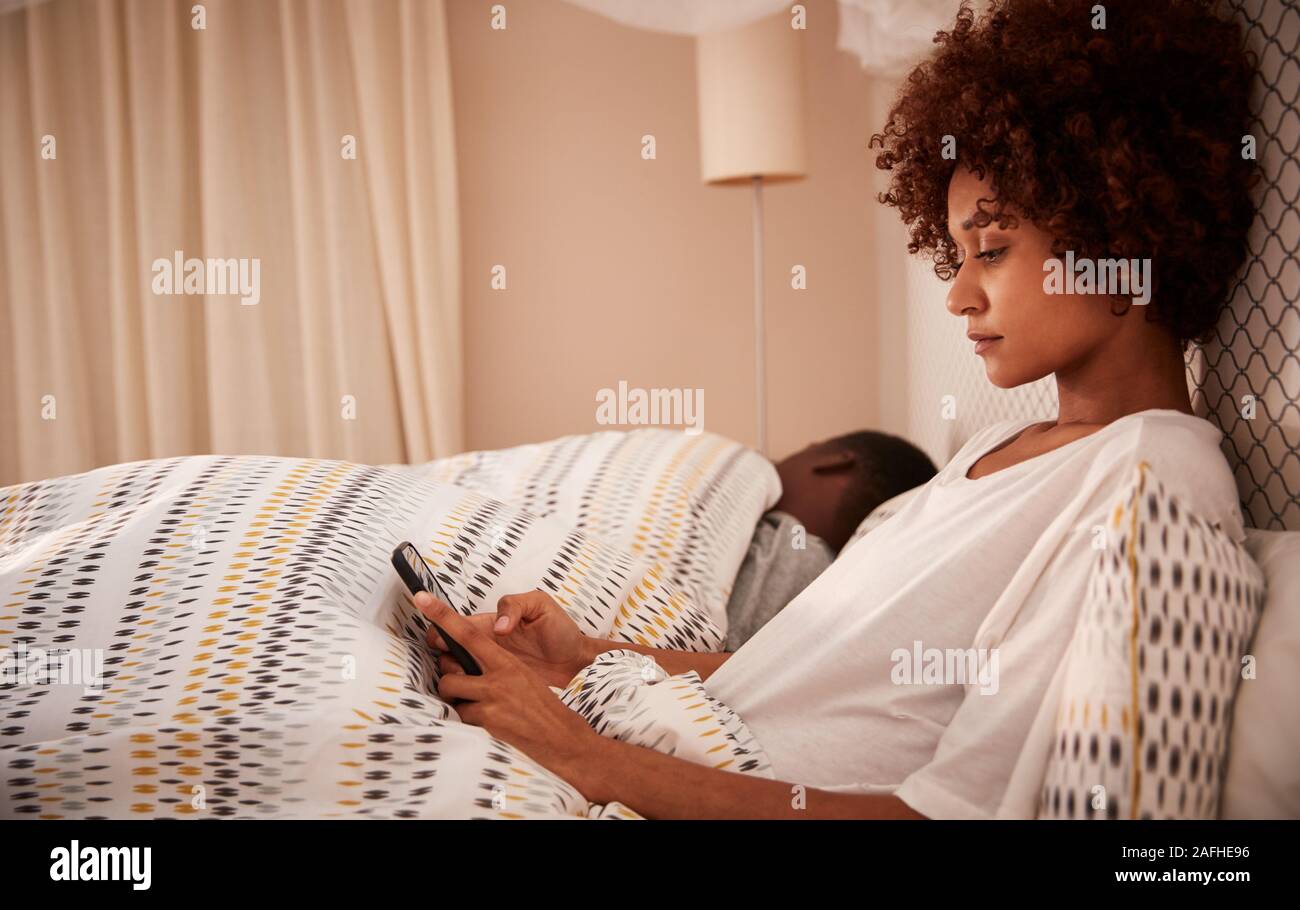 Millennial African American woman sitting up in bed using her smartphone, side view, close up Stock Photo