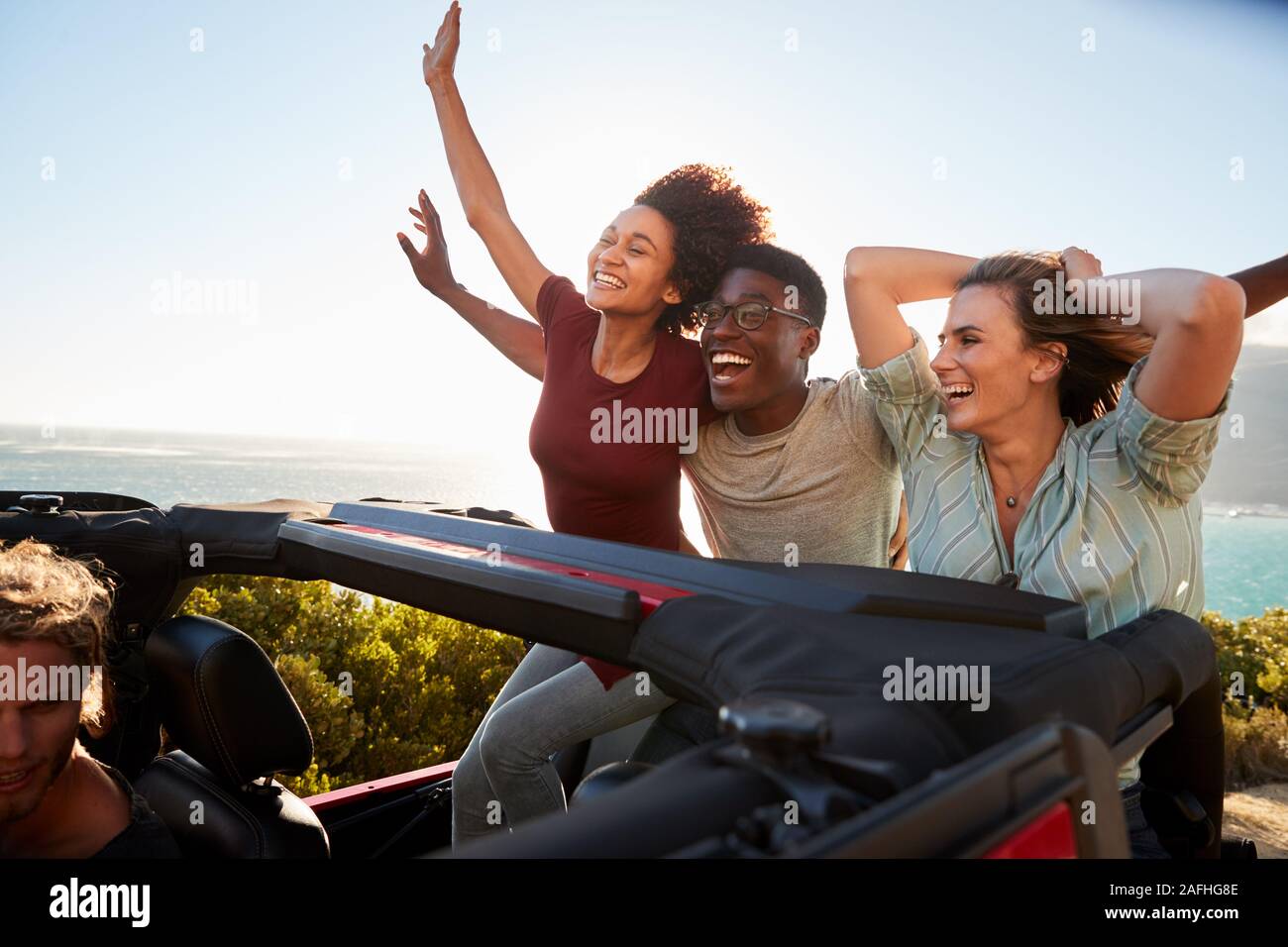 Excited millennial friends travelling in the back of an open car with their arms in the air Stock Photo